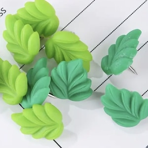 May include: Green leaf-shaped push pins with a silver pin. The pins are arranged on a white surface with black lines.