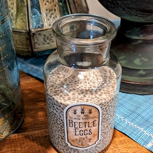 May include: A clear glass jar filled with small, dark brown seeds. The jar has a white label with the text "EST 1989 DELIRIUM APOTHECARY BEETLE EGGS".
