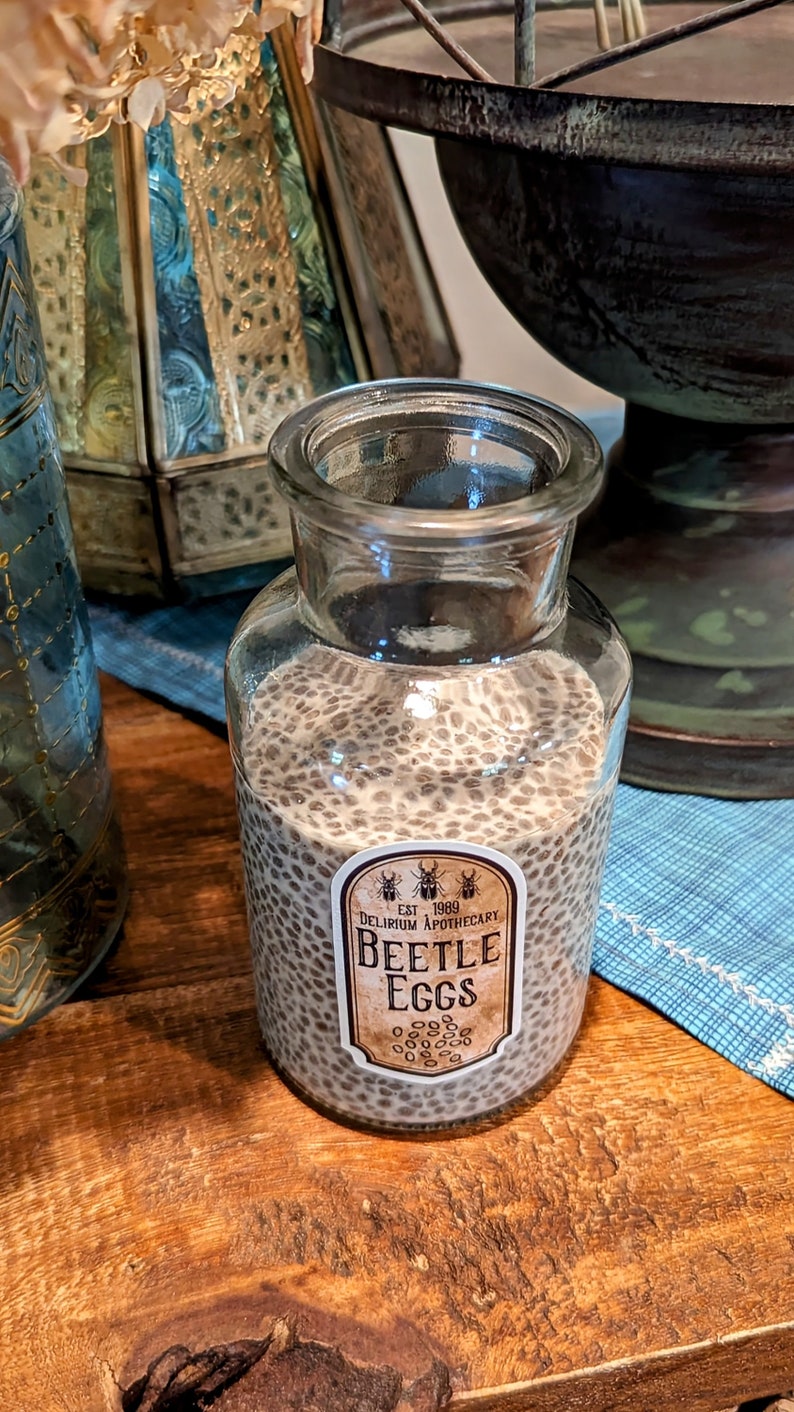 May include: A clear glass jar filled with small, dark brown seeds. The jar has a white label with the text "EST 1989 DELIRIUM APOTHECARY BEETLE EGGS".