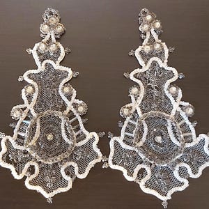 May include: Two white lace appliques with silver beading and sequins. The appliques are shaped like a teardrop and have a floral design.