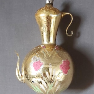 May include: A vintage gold-coloured Christmas ornament shaped like a teapot. The ornament has a round base and a tall, slender neck with a handle and spout. Pink floral designs and green leaves decorate the surface. A silver metal loop is at the top for hanging.