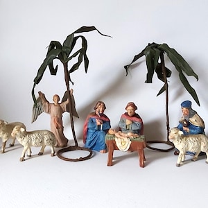 May include: A nativity scene featuring a baby Jesus, Mary, Joseph, an angel, two sheep, and two palm trees. The figures are made of painted plaster and the trees are made of dried leaves.