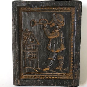 May include: A dark wood carving of a person playing a trumpet in front of a house. The carving is framed in a dark wood frame with a gold border.