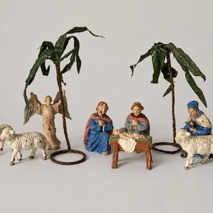 Peut inclure: Une crèche de Noël composée de figurines peintes représentant un ange, un couple central, un bébé dans une mangeoire et trois moutons. Deux grands palmiers en fil de fer brun avec des feuilles en papier vert reposent sur des bases circulaires.