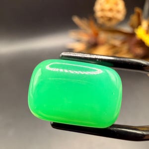 May include: A bright green, rectangular gemstone with rounded corners, held by black tweezers. The polished stone has a smooth, reflective surface. The background is dark, accentuating the vivid colour of the gem.