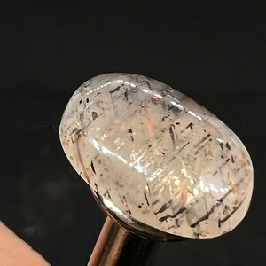 May include: A clear, oval-shaped gemstone with black and silver inclusions. The gemstone is mounted on a silver metal rod.