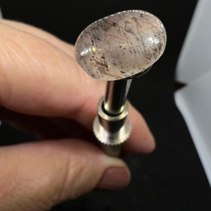 May include: A clear, oval-shaped gemstone with black and brown inclusions, mounted on a silver-colored metal setting.
