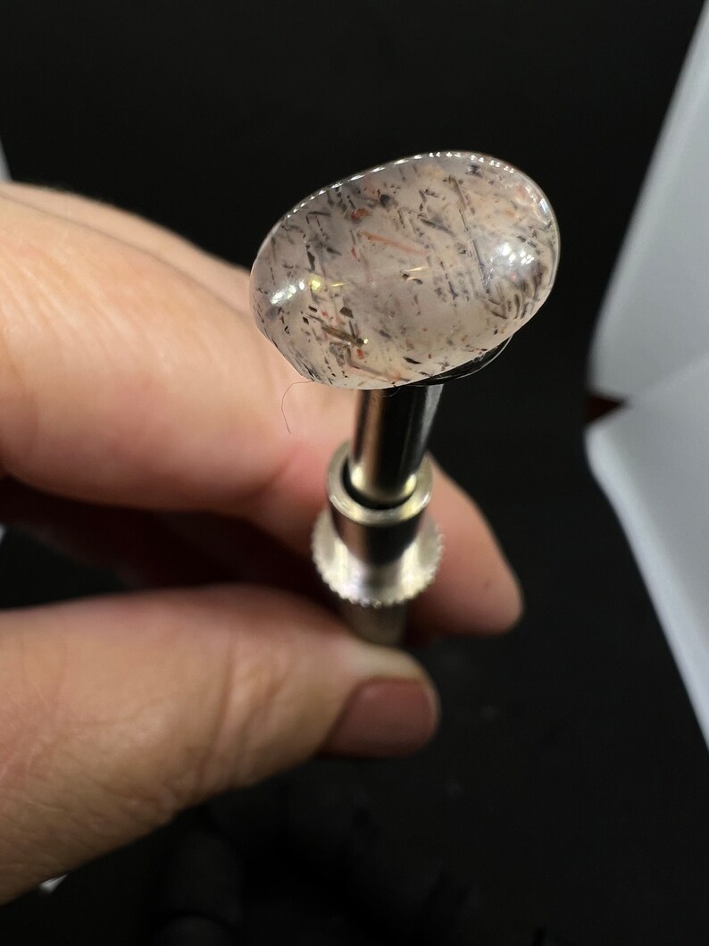 May include: A clear, oval-shaped gemstone with black and brown inclusions, mounted on a silver-colored metal setting.