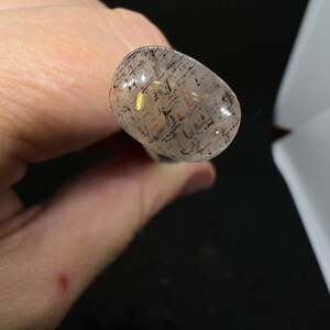 May include: A clear, oval-shaped stone with black and gold flecks. The stone is translucent and has a smooth surface.