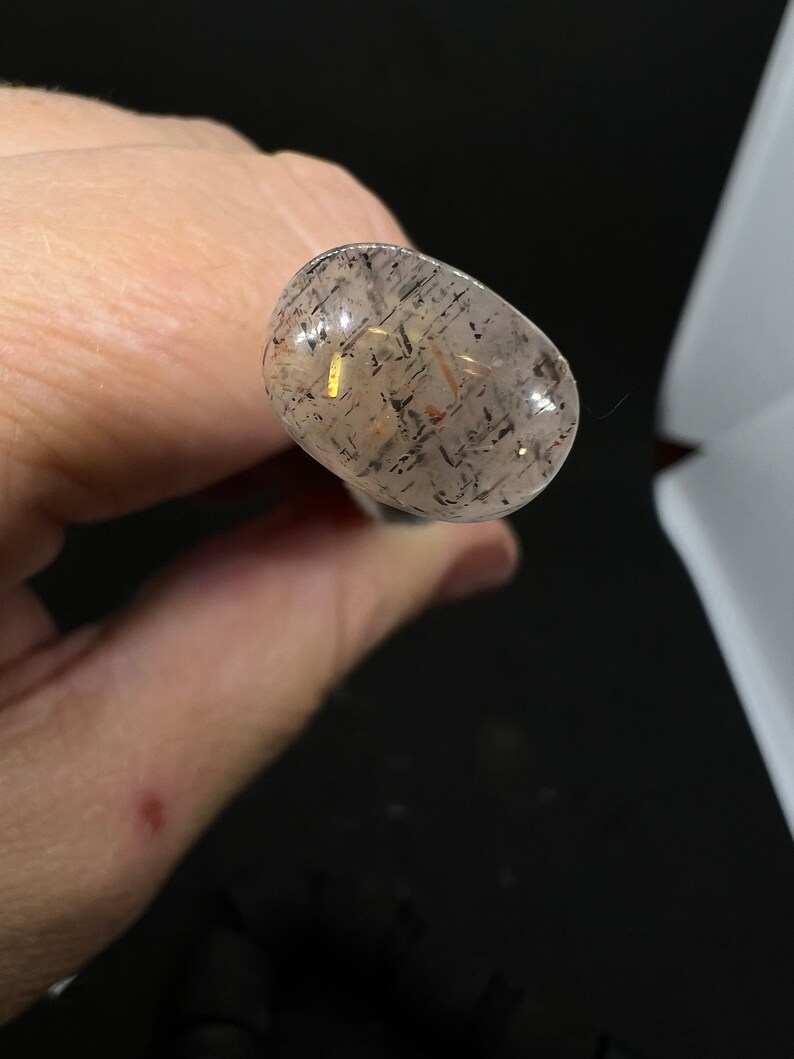 May include: A clear, oval-shaped stone with black and gold flecks. The stone is translucent and has a smooth surface.
