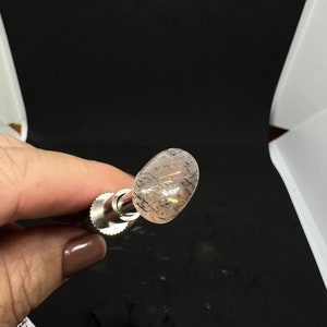 May include: A close-up of a clear, oval-shaped crystal with a pink hue and black inclusions. The crystal is mounted on a silver metal rod.