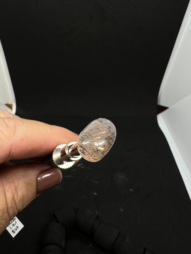 May include: A close-up of a clear, oval-shaped crystal with a pink hue and black inclusions. The crystal is mounted on a silver metal rod.
