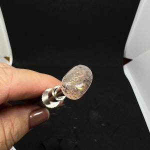 May include: A clear, oval-shaped crystal with a silver metal rod attached to it. The crystal has a subtle shimmer and a few small black inclusions.