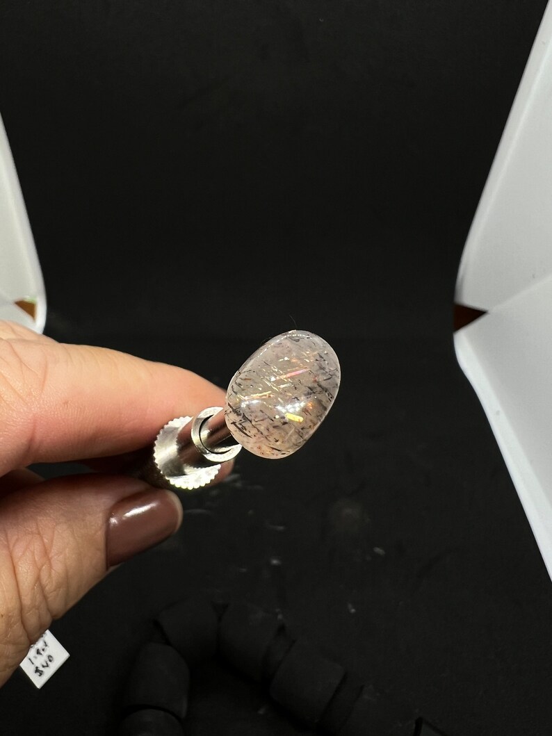 May include: A clear, oval-shaped crystal with a silver metal rod attached to it. The crystal has a subtle shimmer and a few small black inclusions.