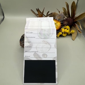 May include: A white gemstone information card with lines for ID Code, Gemstone, Origin, Carats, Measurement, and Price. The bottom section is a black rectangle. The card is in front of dried flowers.