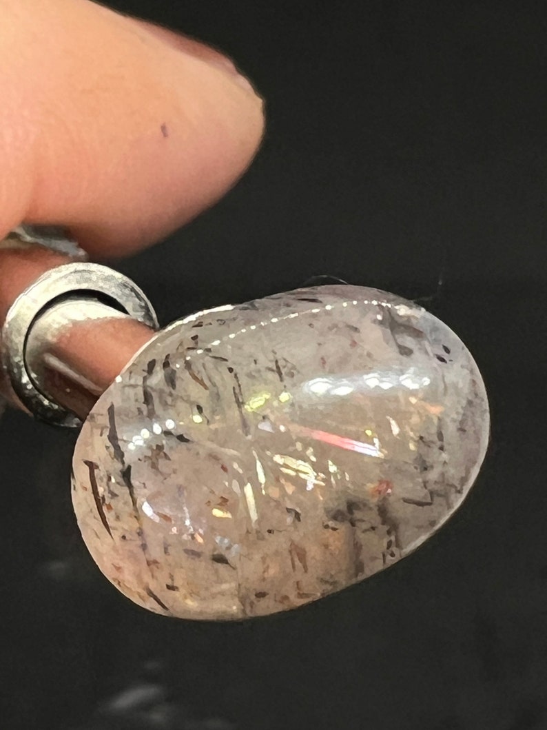 May include: A clear, oval-shaped gemstone with black inclusions. The gemstone is set in a silver setting and is mounted on a silver post.