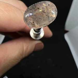 May include: A clear oval-shaped gemstone with black and brown inclusions. The gemstone is held in a metal setting.