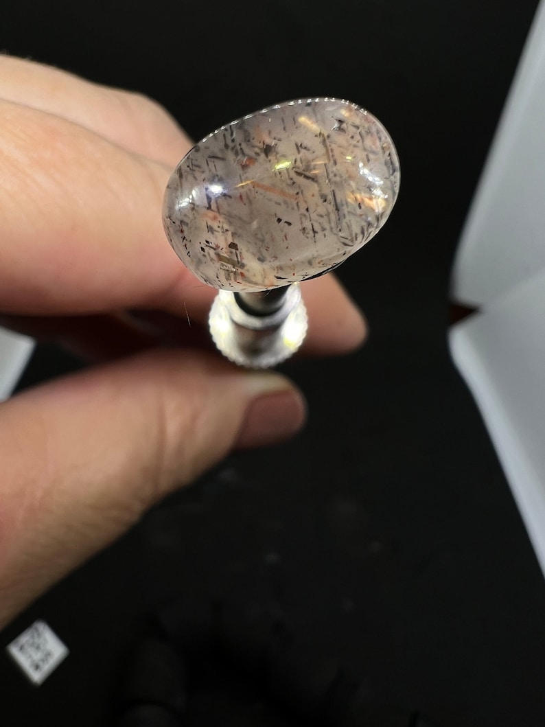 May include: A clear oval-shaped gemstone with black and brown inclusions. The gemstone is held in a metal setting.