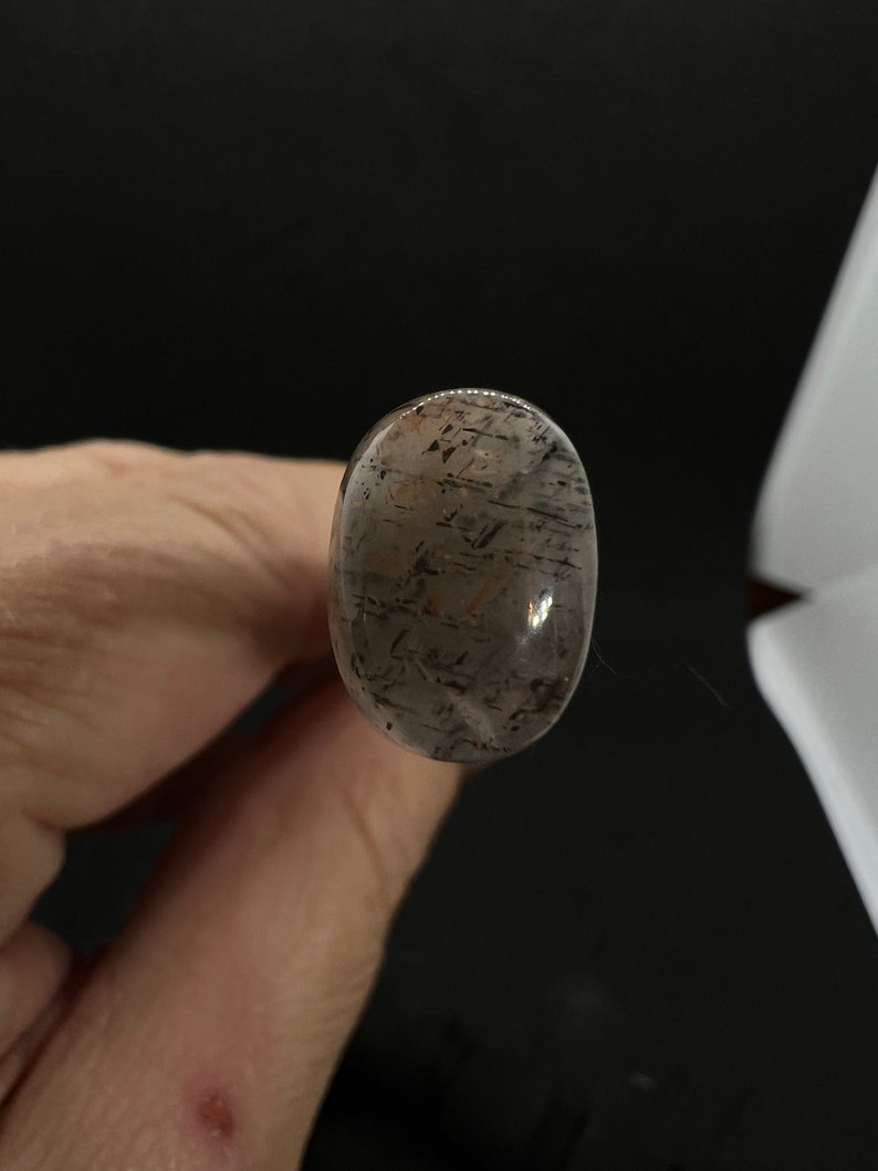 May include: A clear oval-shaped gemstone with black and brown inclusions. The gemstone is translucent and has a smooth, polished surface.