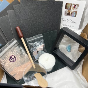 May include: A craft kit containing various items for lapidary work. Includes sandpaper, a wooden tool, small stones, a candle, a small dish with a wooden spoon, and a framed stone. The kit is packaged in a cardboard box.