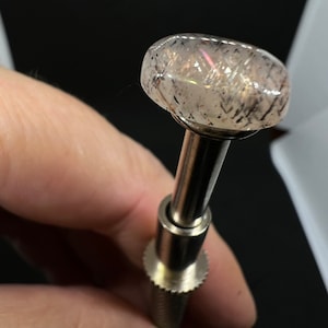 May include: A clear gemstone with black inclusions, set on a silver metal mounting. The gemstone is round and has a slight iridescent sheen.