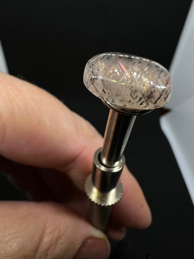May include: A clear gemstone with black inclusions, set on a silver metal mounting. The gemstone is round and has a slight iridescent sheen.