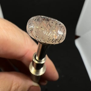 May include: A clear, oval-shaped gemstone with a light pink hue and black and silver flecks. The gemstone is mounted on a silver metal tool.