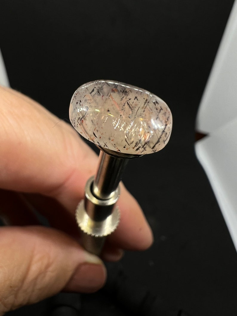 May include: A clear, oval-shaped gemstone with a light pink hue and black and silver flecks. The gemstone is mounted on a silver metal tool.