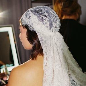 May include: A white lace bridal veil, with detailed floral patterns, covering the head and shoulders. The bride is seen from the back, with dark hair and a bare shoulder. The background is blurred, suggesting a preparation setting.
