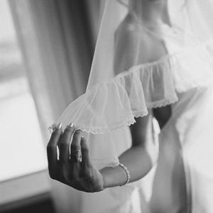 May include: A black and white image of a bride wearing a white dress and veil. The veil is sheer and delicate, with a ruffled edge. The bride's hand is visible, adorned with a ring and bracelet.