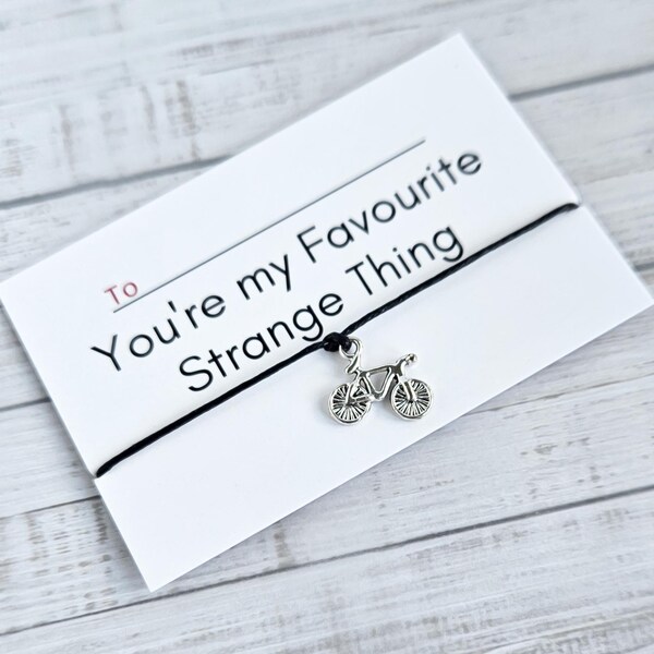 May include: A black string bracelet with a silver bicycle charm and a card that reads "To ____ You're my Favourite Strange Thing".