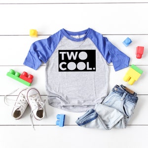 Może przedstawiać: Szara i niebieska koszulka bejsbolowa dla małego dziecka z nadrukiem "TWO COOL." w kolorze czarnym. Koszulka leży na białym drewnianym tle z niebieskimi, czerwonymi i żółtymi klockami.