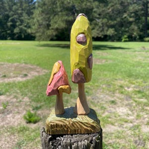 May include: Two wooden mushroom sculptures. One is tall and green with brown accents, the other is smaller and pink with a yellow cap. Both are carved and stand on a wooden base, set against a blurred green background.