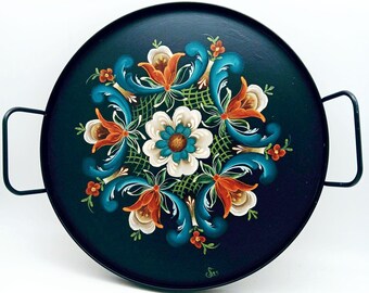 Op de afbeelding: Een ronde zwarte metalen dienblad met een bloemmotief geschilderd in tinten blauw, oranje, wit en groen. Het dienblad heeft twee metalen handvatten.