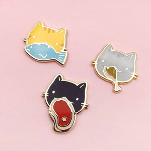 May include: Three enamel pins featuring cartoon cats with different expressions. The first cat is yellow and eating a blue fish. The second cat is gray and eating a brown drumstick. The third cat is black and eating a red steak.