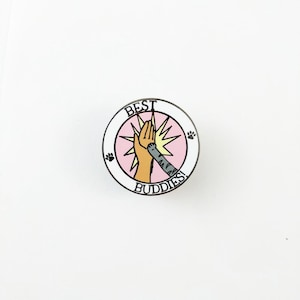 Könnte beinhalten: Eine weiße Emaille-Anstecknadel mit rosa und gelbem Hintergrund. Die Anstecknadel zeigt eine Katze und eine menschliche Hand, die sich gegenseitig ein High Five geben. Der Text "BEST BUDDIES!" ist um den Rand der Anstecknadel herum geschrieben.