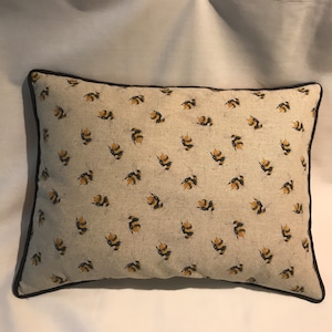 May include: A rectangular throw pillow with a beige linen fabric cover. The pillow has a black trim and a repeating pattern of yellow and black bumblebees.