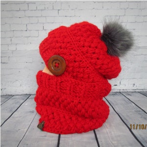 May include: Red crocheted hat and neck warmer set with a faux fur pom pom and a wooden button. The set is photographed on a wooden surface.