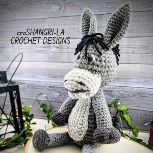 Könnte beinhalten: Ein grauer gehäkelter Esel mit schwarzem Garn für Mähne und Schweif. Der Esel sitzt auf einer Holzscheibe. Der Text "croSHANGRI-LA CROCHET DESIGNS" ist im Hintergrund sichtbar.