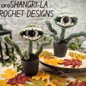 May include: Two crocheted green plant-like creatures with white teeth and a single black eye. The creatures are in black pots and sitting on a wooden slice with fall leaves.