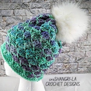 May include: A hand-crocheted beanie hat in teal, green, and purple, featuring a large white faux fur pom-pom. The hat is displayed on a white mannequin head. The text "croSHANGRI-LA CROCHET DESIGNS" is visible.