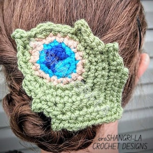 以下が含まれることがあります： 青と茶色の目を持つ緑色のクロシェの孔雀の羽根のヘアクリップ。クリップは女性の茶色の髪につけられています。