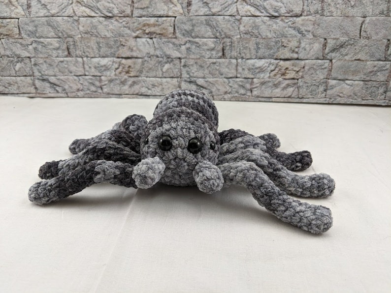 K&ouml;nnte beinhalten: Ein graues, geh&auml;keltes Spinnen-Pl&uuml;schtier mit schwarzen Augen. Die Spinne hat acht Beine und liegt auf einer wei&szlig;en Oberfl&auml;che.