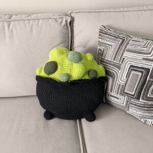 K&ouml;nnte beinhalten: Ein geh&auml;keltes Kesselkissen mit einem gr&uuml;n-schwarzen Design. Der Kessel ist mit einem gr&uuml;n-braunen, geh&auml;kelten Brei gef&uuml;llt. Das Kissen liegt auf einem hellgrauen Sofa.