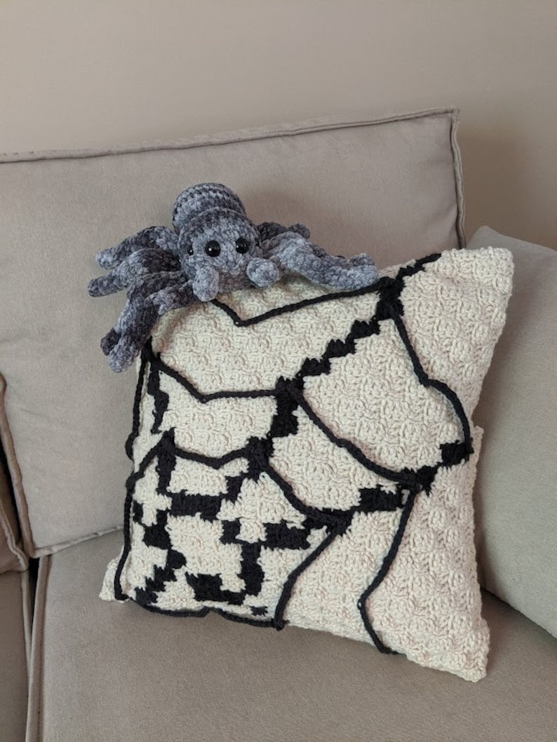 K&ouml;nnte beinhalten: Ein geh&auml;keltes Kissen mit einem schwarzen und wei&szlig;en Spinnennetz-Design. Eine pl&uuml;schige graue Spinne sitzt auf dem Kissen.