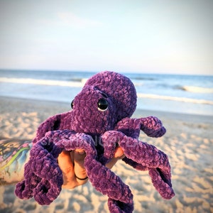 May include: Purple crocheted octopus plush toy with black eyes. The octopus is being held in a hand against a beach background.