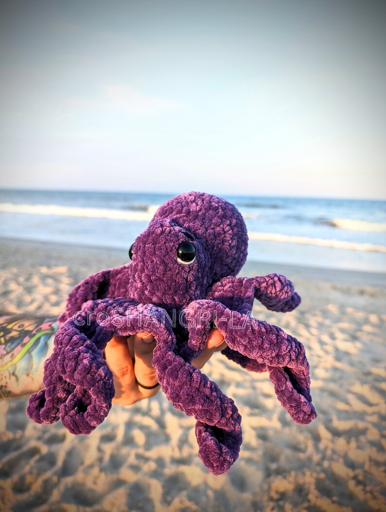 May include: Purple crocheted octopus plush toy with black eyes. The octopus is being held in a hand against a beach background.