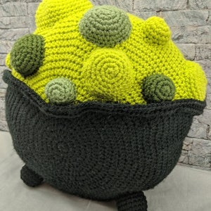 K&ouml;nnte beinhalten: Ein geh&auml;keltes Kesselkissen mit einem gr&uuml;n-gelben Design. Der Kessel hat drei Beine und ist mit gr&uuml;nen und gelben Kugeln gef&uuml;llt.