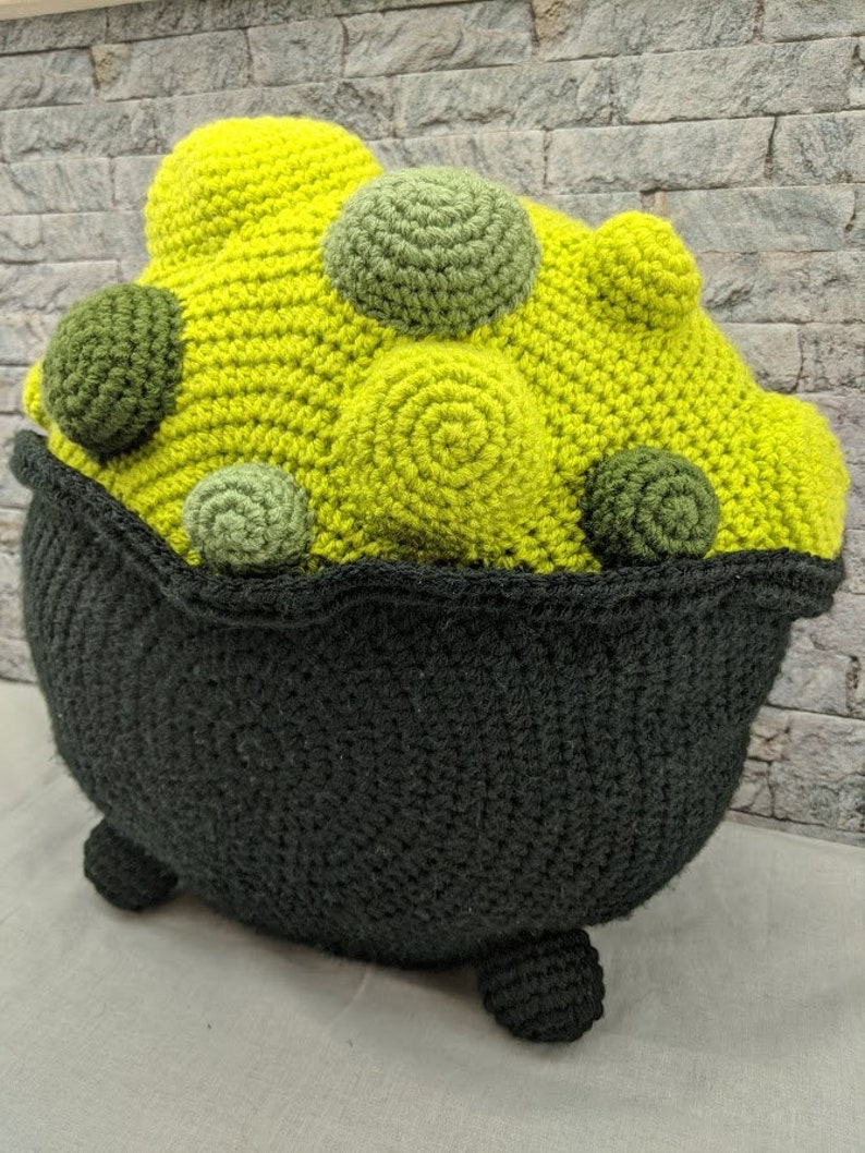 K&ouml;nnte beinhalten: Ein geh&auml;keltes Kesselkissen mit einem gr&uuml;n-gelben Design. Der Kessel hat drei Beine und ist mit gr&uuml;nen und gelben Kugeln gef&uuml;llt.