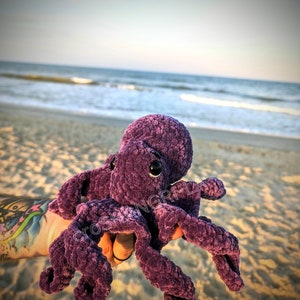 May include: A purple crocheted octopus plush toy with large black eyes. The toy is being held by a hand with a tattoo on the beach.